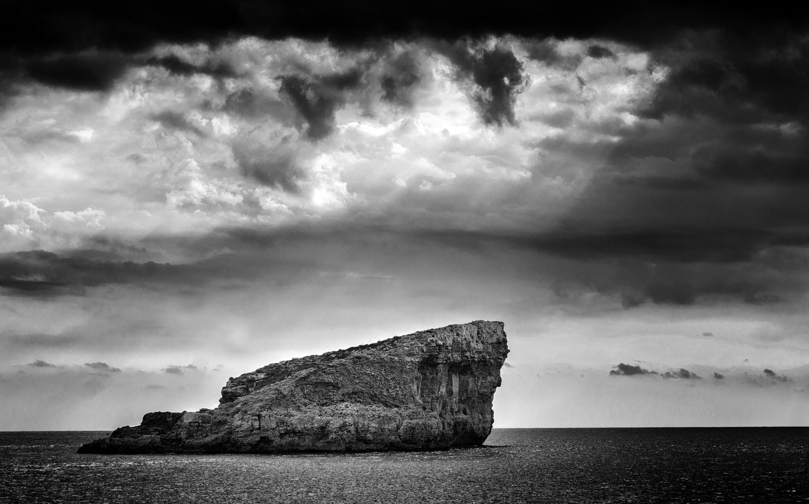 A bare rock islet off Comino under heavy clouds with rays of light, black and white