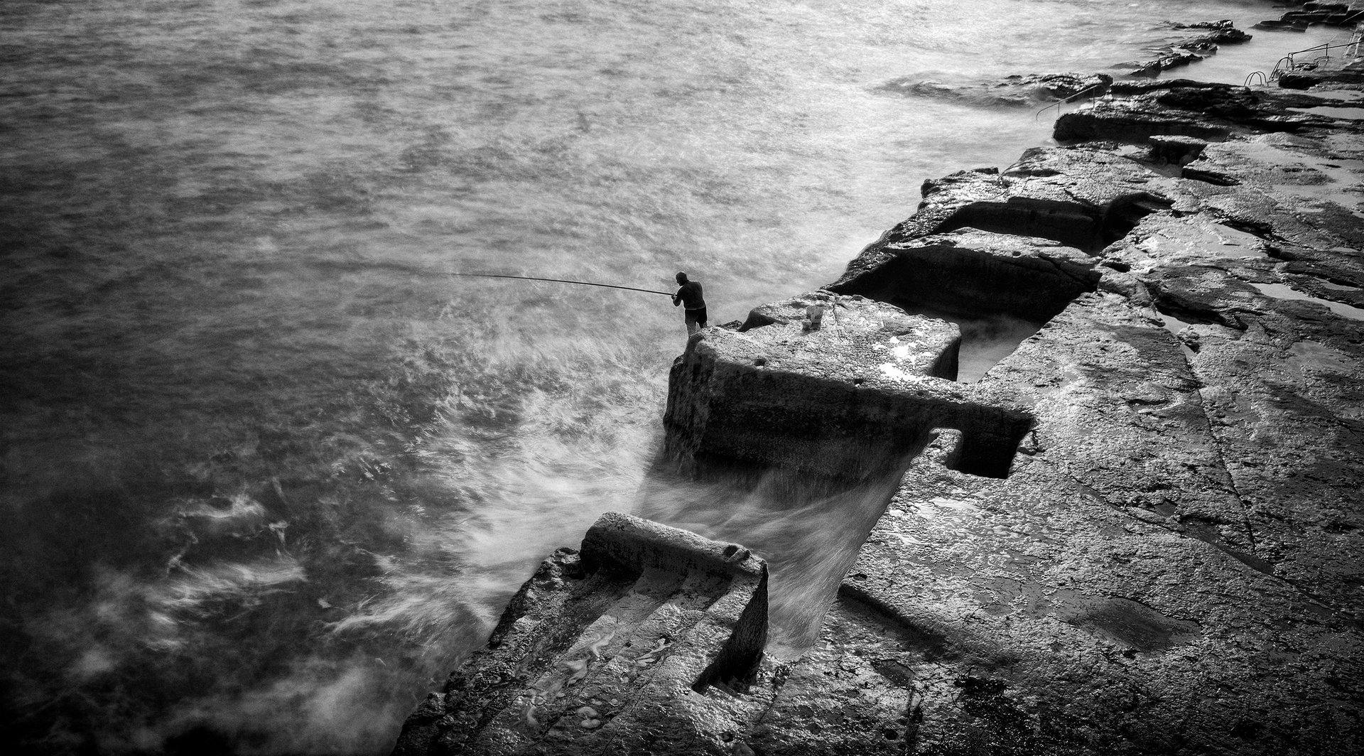 A lone fisherman on stone shelves stepping into a rough sea at Sliema, black and white