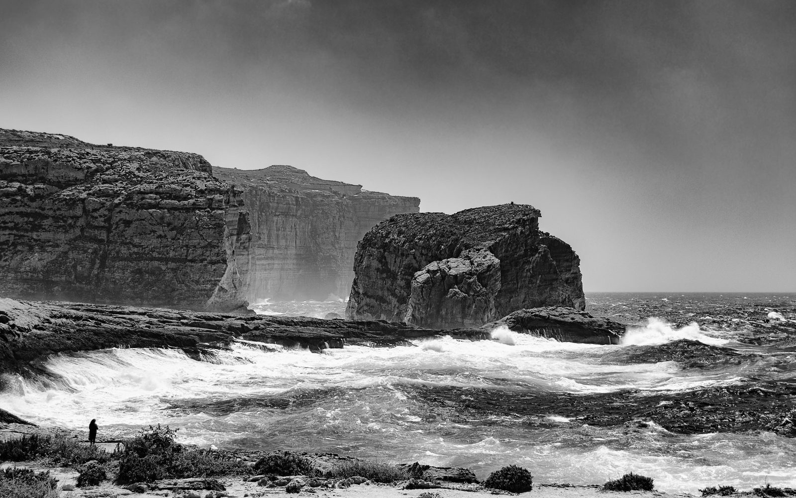 A lone figure on the shore facing Ġebla tal-Ġeneral rock, black and white