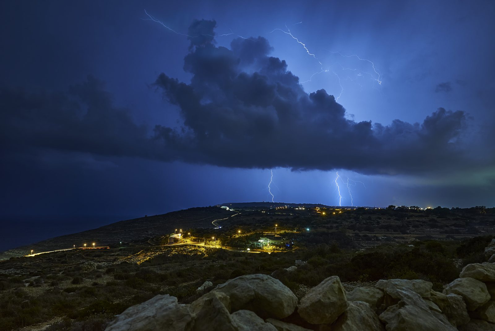 Three lightning bolts striking over the lit hillside at Żurrieq, Malta